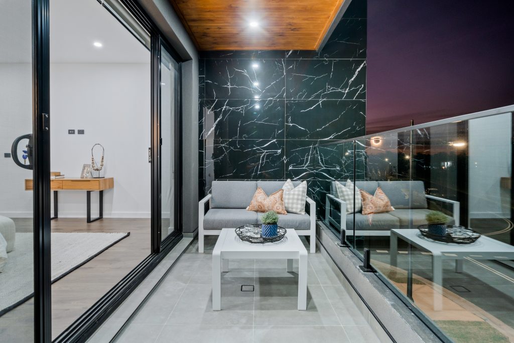 Image shows a spacious balcony with grey tiled floor and black granite tiled wall with glass railing overlooking a road