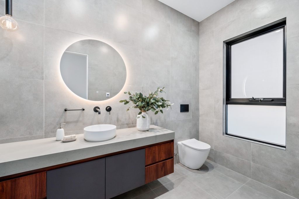 Image shows a bathroom with grey stone tiles floor to ceiling, toilet and sink, well-lit, with a round mirror