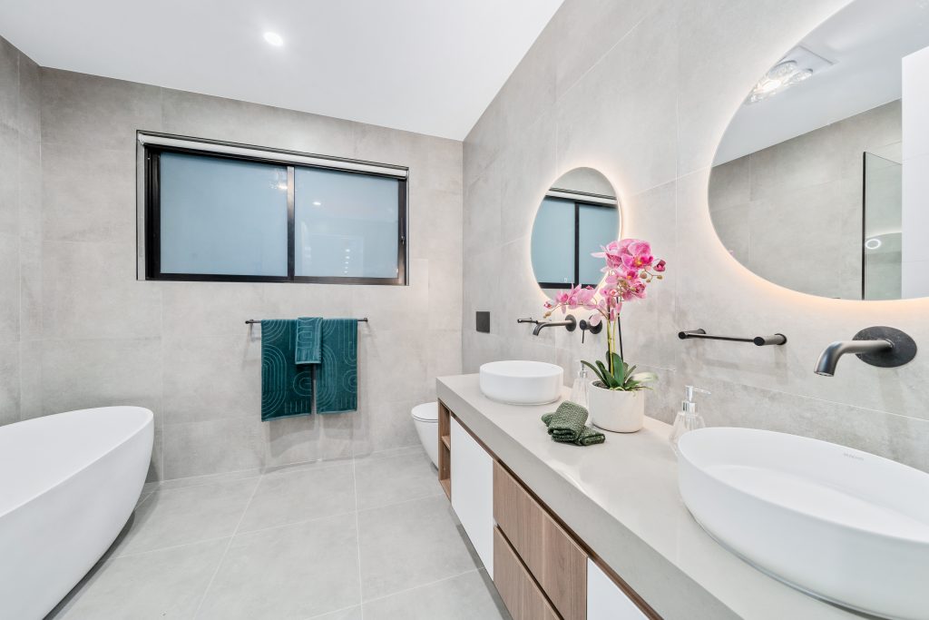 Image shows a bathroom with grey tiles floor to ceiling. Two round mirrors with led lighting. White bath and toilet