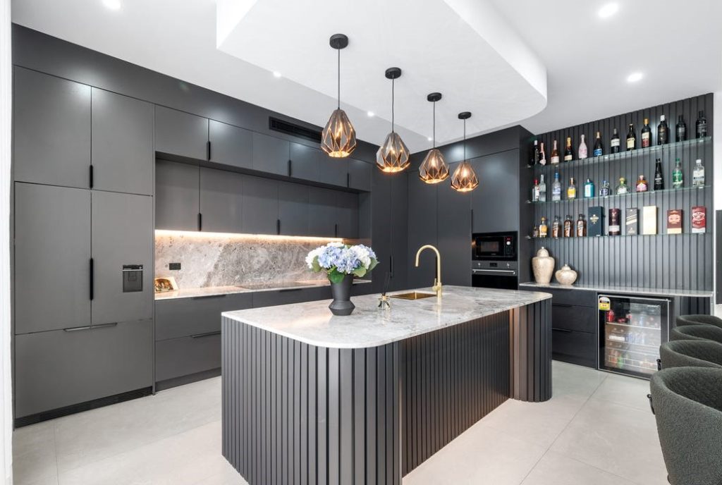 Image shows a modern kitchen with high-end interior design. Custom cabinetry, granite kitchen bench-tops and built-in oven, microwave, and cook tops. Well lit with mood lighting. Designed and built by Sydney based home builders Benelong Projects