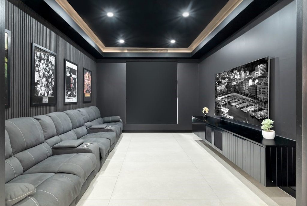 Image shows a home cinema with large TV, dark walls, and comfortable seating. Custom entertainment unit and down-lighting