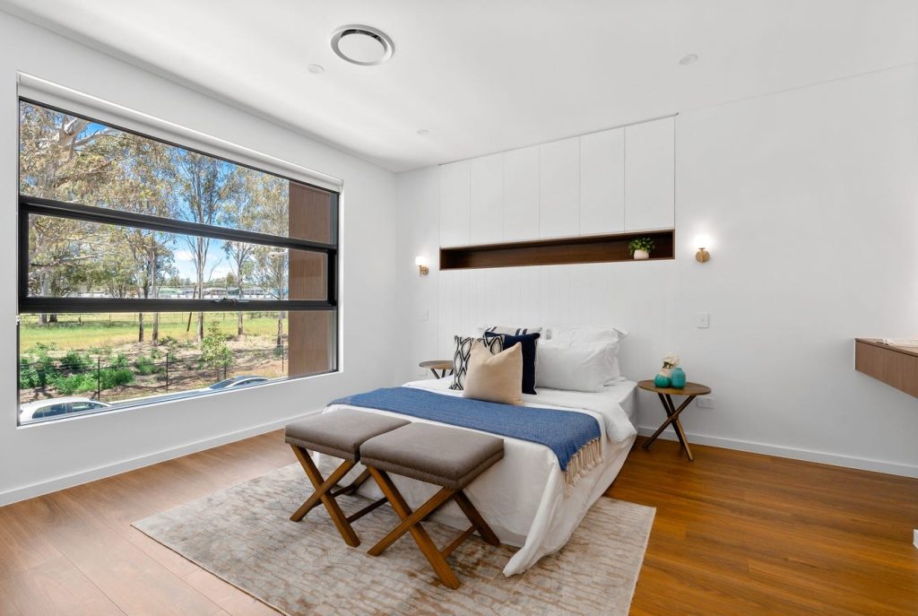 Image shows modern spacious well-lit bedroom. There is a bed with side tables. In-built table for make-up and getting ready. The room is well lit and has wooden floors. Designed and built by Sydney based home builders Benelong Projects