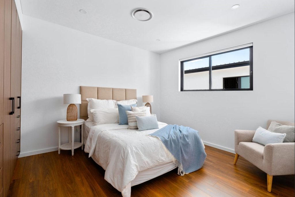 Image shows a large well-lit bedroom with natural light. Wooden floors and custom cupboards. Bed with side table, lamps, and arm chairs. Build and designed by Benelong Projects in Sydney