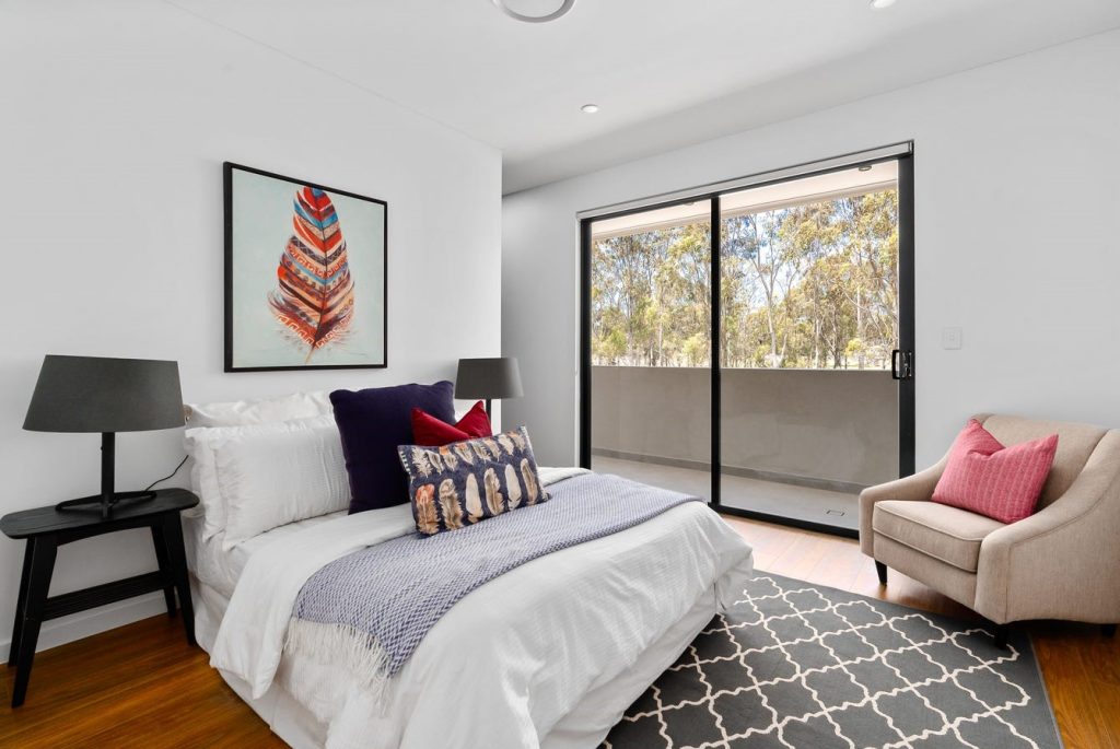 Image shows a spacious bedroom with bed, side table, chairs and lamps. Well lit with natural light with large ranch slider. Wooden floors with a grey rug.