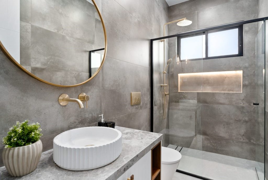 Image shows a bathroom designed and built by Sydney based home builders Benelong products. Grey stone tiles on the floor and ceiling, large shower with gold brass fittings, white sink and toilet. Well lit with mood lighting and indoor plants.