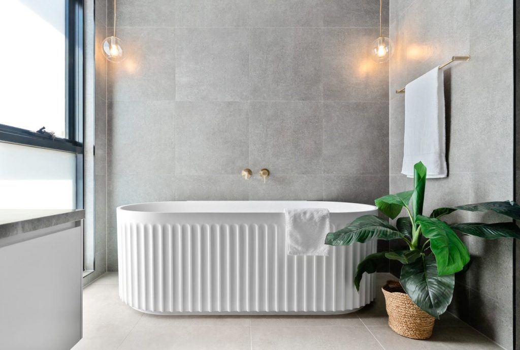 Image shows a white bath in a bathroom with grey stone tiles on the floor and ceiling. Gold coloured towel rain on the wall. Indoor plants with mood lighting. Well lit with natural light.