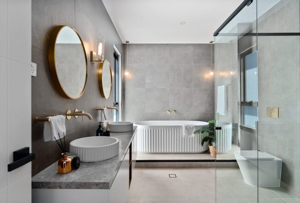 Image shows a large spacious bathroom with stone tiles on the floor and walls. There are two sinks, a bath, toilet, and large shower. Mood lighting. Designed and built by Sydney based home builders Benelong Projects