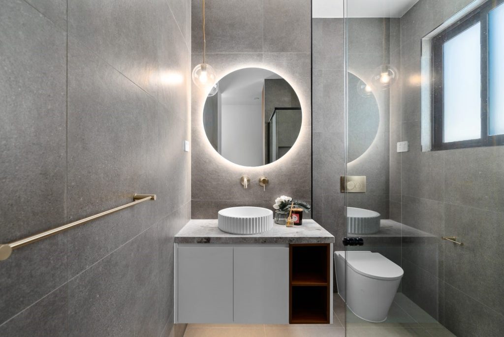 Image shows a bathroom with modern interior design built by Sydney based builders Benelong Projects. The floor and walls feature large stone tiles while the benchtop is grey granite. There is a sink, toilet, and a large spacious shower with brass / gold coloured fittings