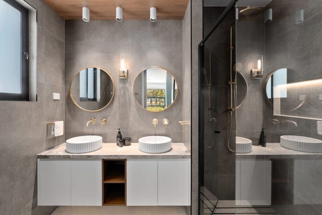 Image shows a bathroom with modern interior design built by Sydney based builders Benelong Projects. The floor and walls feature large stone tiles while the benchtop is grey granite. There are two sinks and a large spacious shower with brass / gold coloured fittings