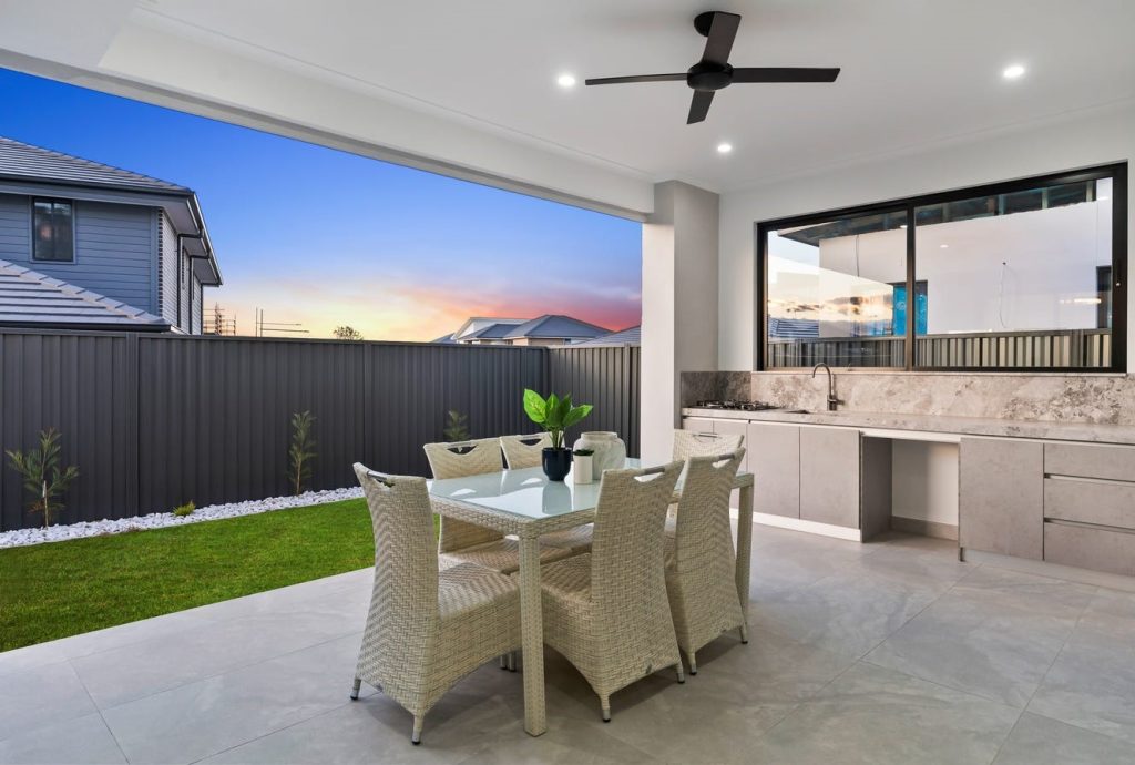 Image shows a spacious outdoor living and dining area complete with cooking area with granite bench-tops, premium fittings and gas stove. The flooring is stone tiled and backs onto as lush garden with plants. Outdoor table and chair set for fine dining