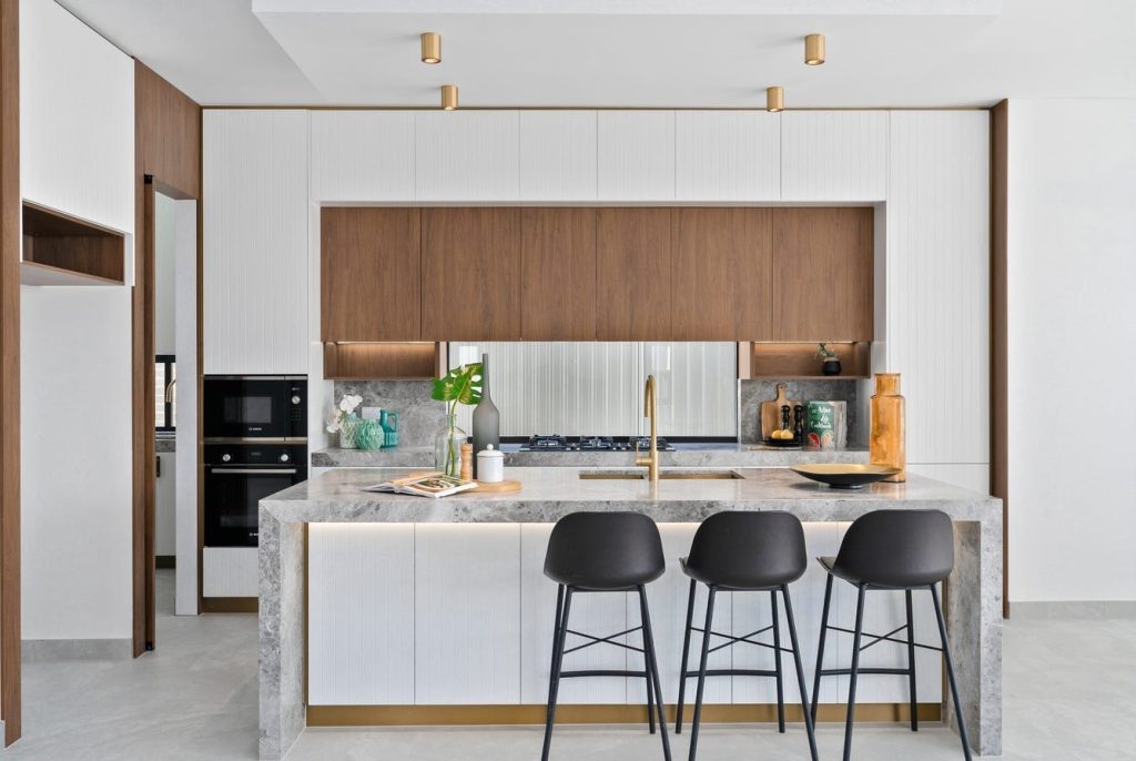 Image shows a kitchen with granite bench-top and stone tile floor. Breakfast bar with stools and granite top. Gas cook-top and in-built microwave and oven. Custom cabinetry makes great use of space. Well-lit