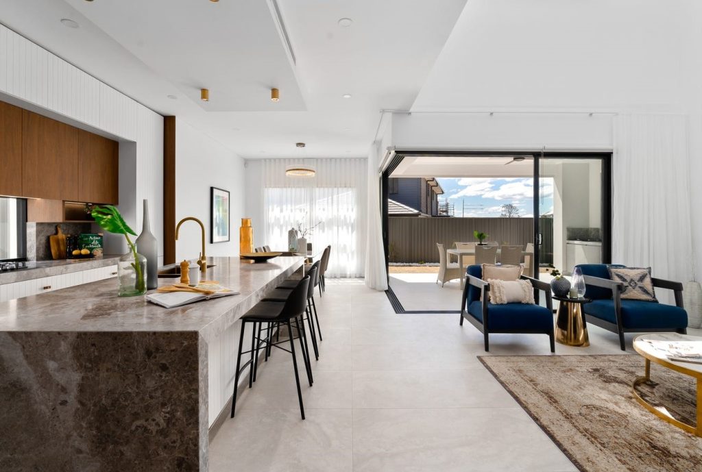 Image shows an open-plan living and dining area backing onto outdoor entertaining area. Grey stone tiles with granite bench-tops and in-built oven and microwave. Blue arm chairs with tables for entertaining. Designed and built by Sydney based home builders Benelong Projects