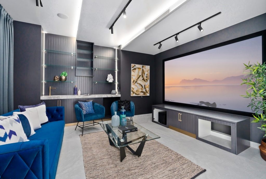 Image shows a home-cinema area built and designed by Sydney based builders Benelong Projects. Large projector TV with cinema-style down-lighting. Comfortable blue chairs for entertaining with glass table.