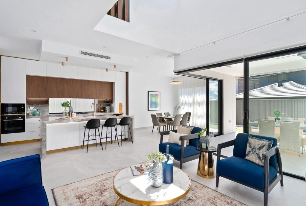 Image shows an open-plan living and dining area backing onto outdoor entertaining area. Grey stone tiles with granite bench-tops and in-built oven and microwave. Blue arm chairs with tables for entertaining. Designed and built by Sydney based home builders Benelong Projects