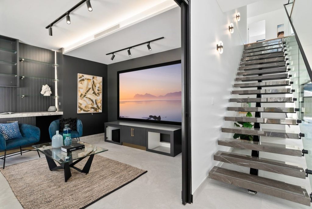 Image shows a spacious entertaining and living area with large projector TV and cinema-style down-lighting. Open-plan with granite staircase and custom storage. Designed and build by Sydney based home builders Benelong Projects