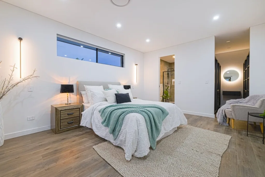 Image shows a white bedroom with large windows, bed, wardrobe, and wooden floors