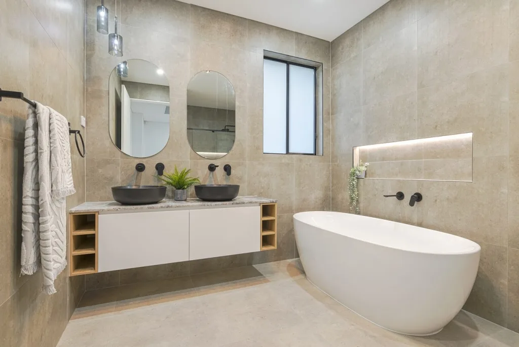 Image shows a large bathroom with bath, hand basins, mirrors, and floor to ceiling tiles