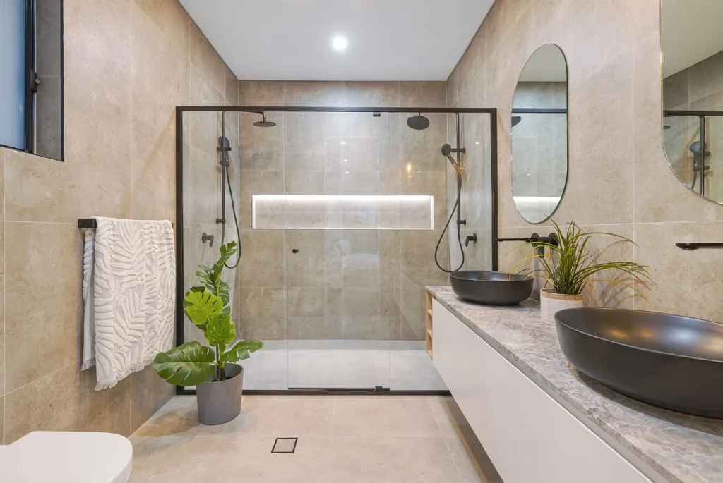Image shows a spacious bathroom with bath, hand basins, mirrors, and sand colored floor to ceiling tiles
