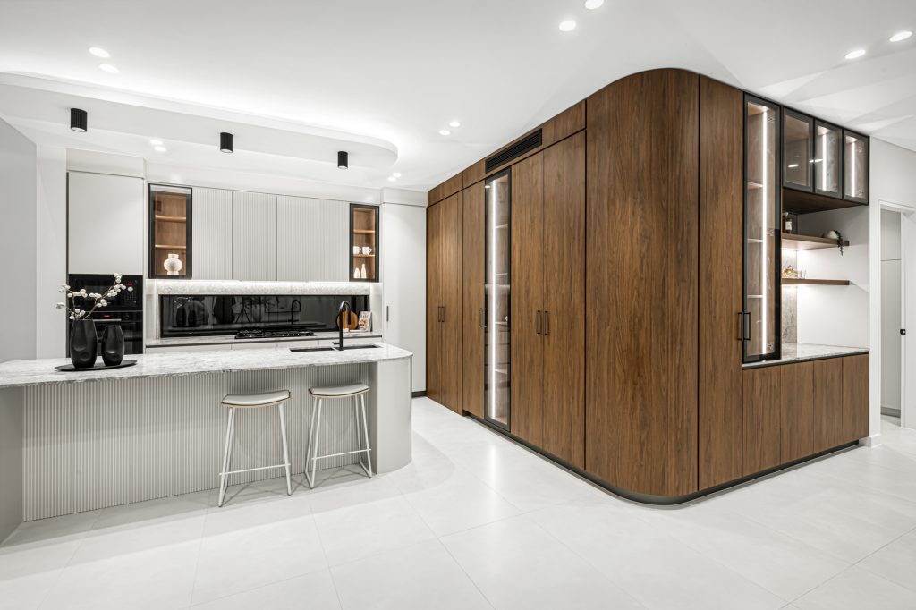 Image shows a modern open plan kitchen area with granite bench-tops and tiled floors. Custom wooden cupboards and storage area designed and built by Benelong Projects, Sydney