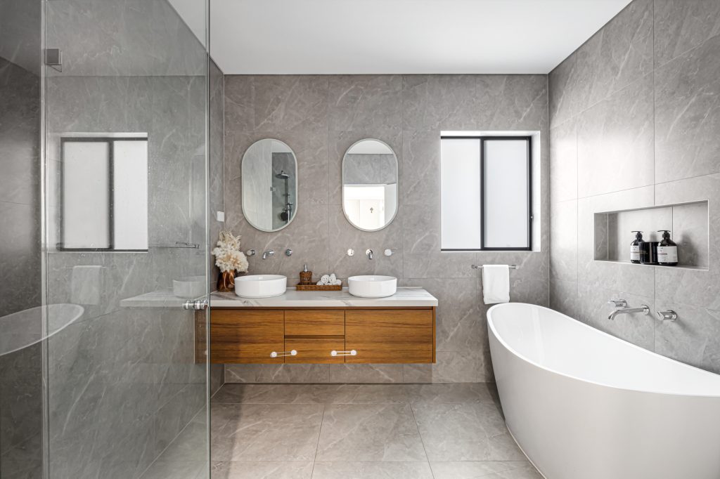 Image shows a large bathroom with double sinks, large shower and bath. Tiled floor to ceiling with grey granite tiles.