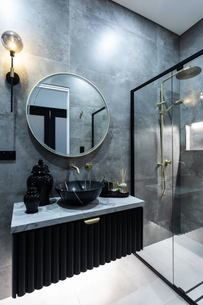 Image shows a bathroom with large shower, dark grey tiled walls, light grey tiled floor and sink with white marble bench top
