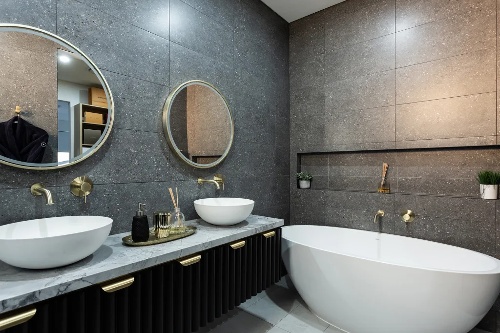 Image shows a stylish and modern bathroom with dark grey tiled walls, light grey floor tiles, a white bath and round mirrors