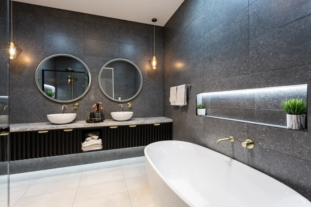 Image shows a bathroom with dark grey tiled walls, light grey floor tiles, a white bath and two round mirrors. Granite bench top with two sinks