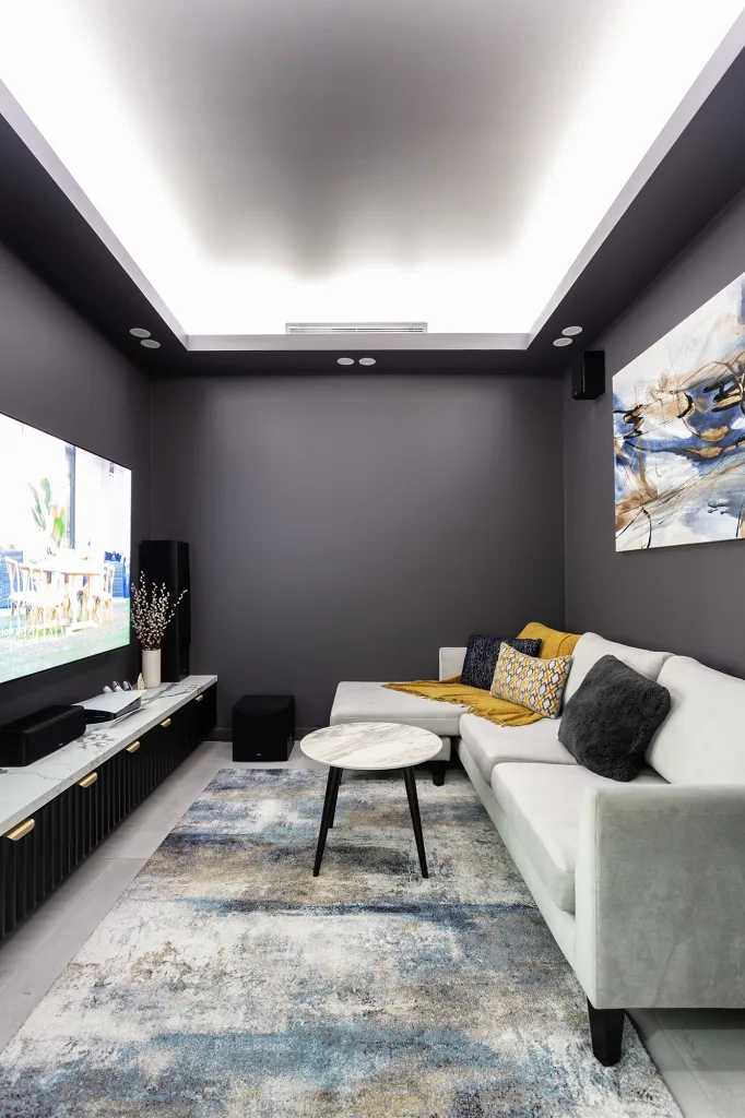 Image shows a home theatre room with dark walls, large TV, and comfortable seating