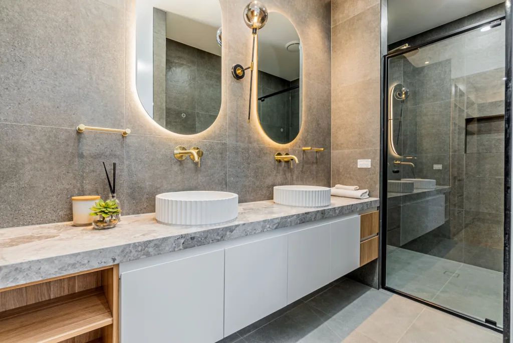 Image shows a modern bathroom with two sinks, grey tiled floors, large shower and double mirrors