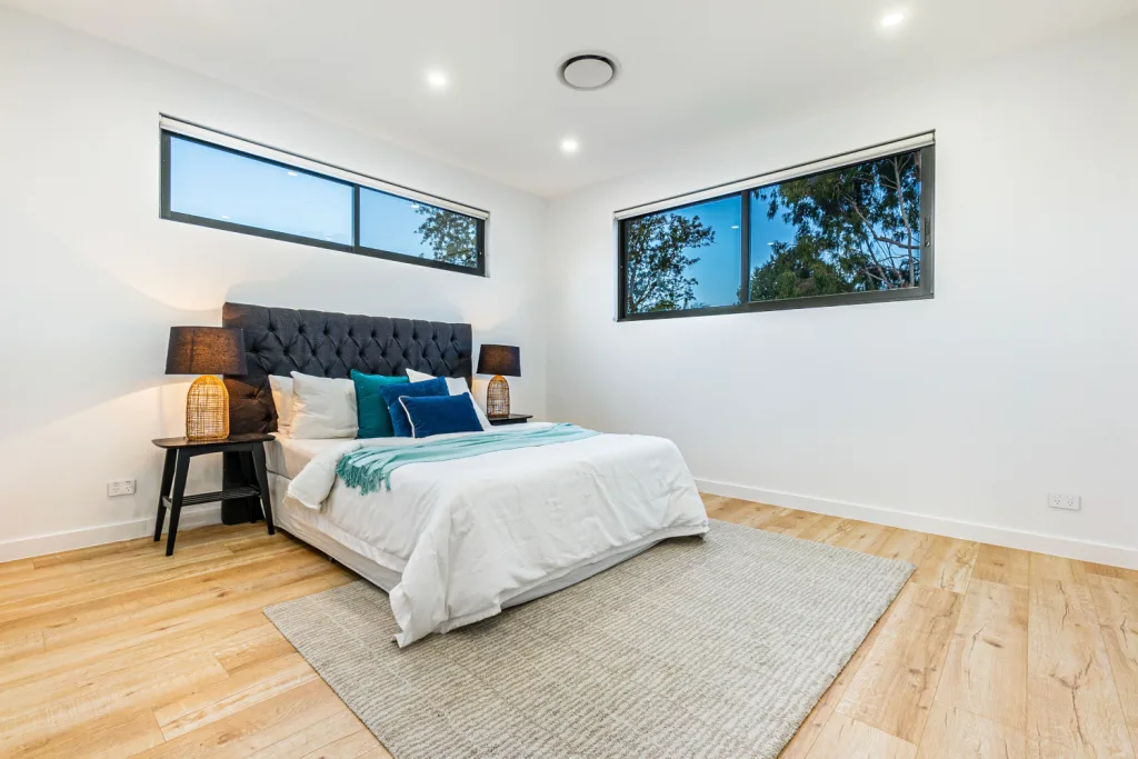Image shows a bedroom with good natural lighting. There are white walls with wooden floors, a large comfortable bed, and a rug on the floor