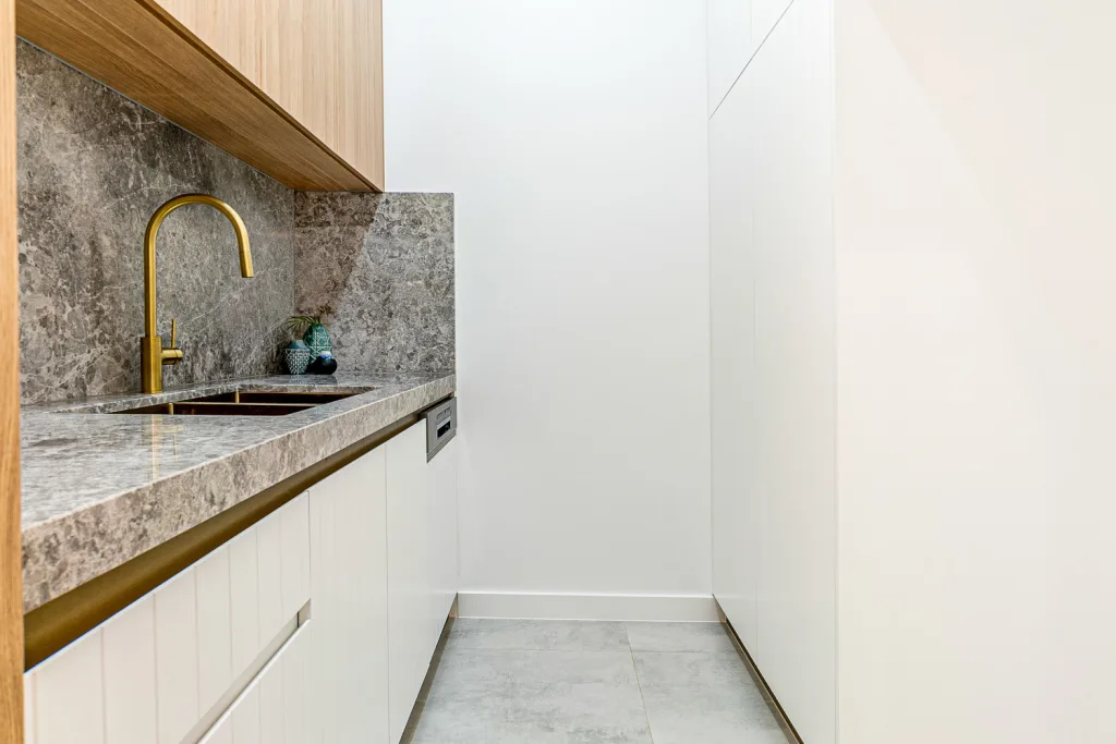 Image shows a luxury laundry with grey granite bench tops, white walls, brass fixtures and grey tiled floor