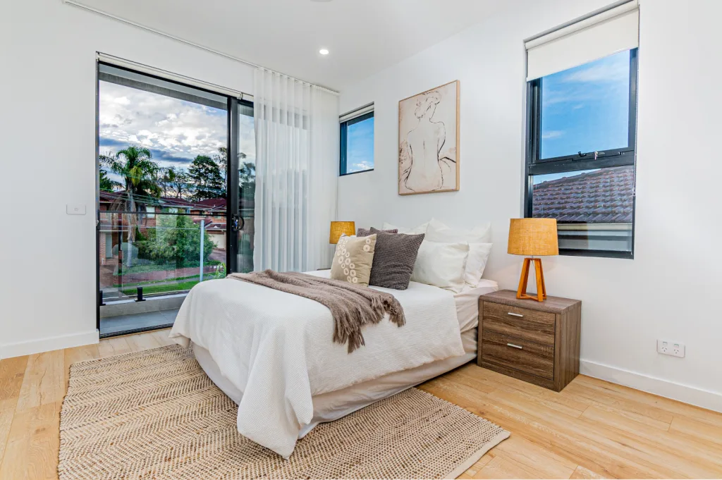 Image shows a bedroom well-lit with natural light. There are white walls with wooden floors